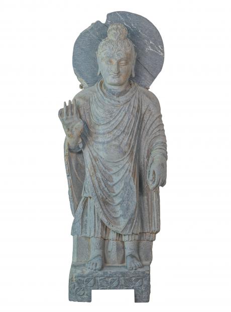 <h3>Buddha in Reassuring Pose</h3> <p>Grey Schist<br>2nd - 3rd century CE<br>Dharmarajika<br>86.5x32x13cm<br>A stone cult image of the haloed Buddha stands on a low pedestal. His right hand is raised in the gesture of reassurance (abhaya-mudra) and the left hand holds the edge of the robe. The gesture represents protection, as the raised right hand symbolizes a shield as well as overcoming fear. The ritual gesture abhaya-mudra is widely prevalent. </p>