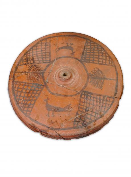 <h3>Lid of the terracotta pot</h3>  <p>A terracotta lid of a storage pot or jar, painted with peacocks, trees and cross-hatching within a circular frame.</p>